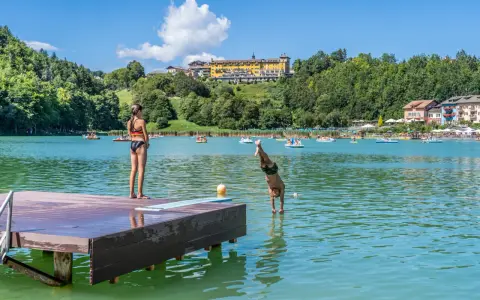 Ragazzo si tuffa in un lago balneabile, mentre una ragazza osserva dal pontile. Altri bagnanti e pedalò popolano le acque.