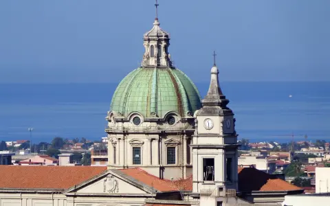 Cupola verde e campanile con orologio a Barcellona Pozzo di Gotto, con il mare azzurro sullo sfondo.