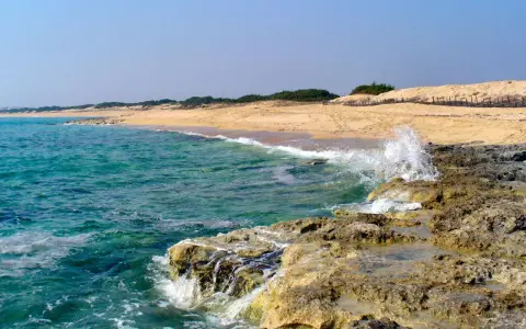 Onde cristalline del mar Ionio in Puglia si infrangono su scogli dorati, con una spiaggia sabbiosa e dune sullo sfondo.