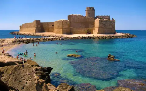 Castello Aragonese sull'acqua cristallina della costa ionica Calabria, con bagnanti che si godono il sole e il mare.