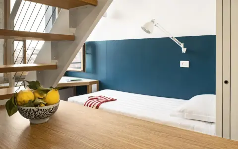 Un letto singolo con lenzuola a righe e un cuscino bianco, sotto una scala in legno e metallo. Un ciotola di limoni sul tavolo crea un'atmosfera da combo Venezia.