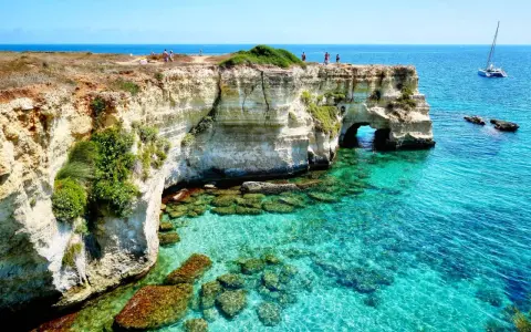 Scogliere bianche e mare cristallino sul litorale adriatico. Turisti ammirano il panorama, un catamarano naviga in lontananza.