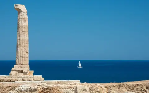 Antica colonna sulla costa degli Achei, con un veliero solitario all'orizzonte sotto un cielo azzurro.