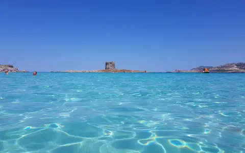 Acque cristalline e un antico torrione su un'isola: un mare bello in Italia, con gente che nuota e una barca a vela all'orizzonte.