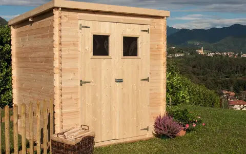La casetta in campagna, in legno chiaro, con due finestrelle e un cesto di legna, si affaccia su un panorama collinare con un borgo in lontananza.