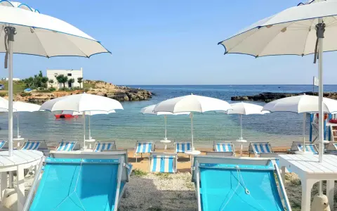 Lido Cala Pescatore: lettini azzurri e ombrelloni bianchi su una spiaggia di ciottoli, con il mare cristallino e rocce sullo sfondo.