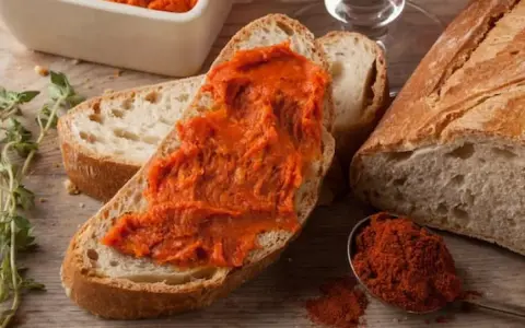 Fetta di pane con 'nduja, uno dei piatti tipici della Calabria, accanto a una ciotola con la crema e un cucchiaino di peperoncino in polvere.