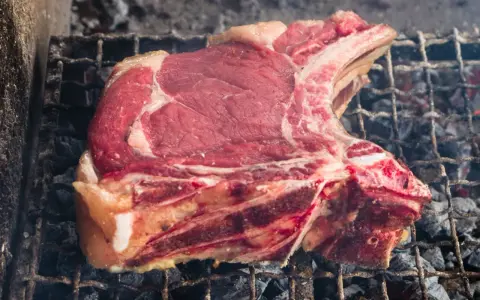 Una succulenta fiorentina di vitello, con la sua succosa carne rossa e il grasso invitante, cuoce lentamente sulla griglia ardente, sprigionando un profumo delizioso.