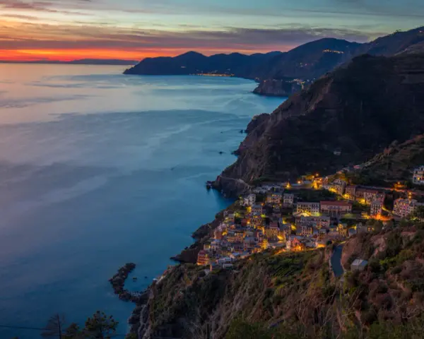 Riomaggiore cosa vedere: un villaggio colorato aggrappato alla scogliera, illuminato al tramonto, con il mare blu e le montagne sullo sfondo.