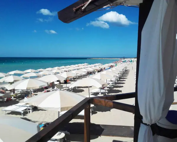 Ombrelloni bianchi e lettini ordinati sulla sabbia dorata, con il mare azzurro e il cielo limpido a fare da sfondo. Un'immagine perfetta di mamire lido Marini.