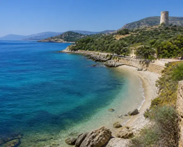 Spiaggia di Guidaloca con acque cristalline, vegetazione mediterranea e una torre antica in collina.