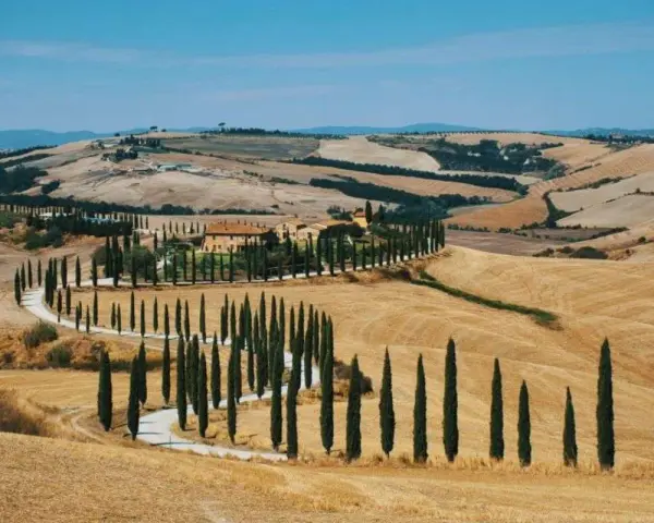 Un casale toscano circondato da cipressi lungo le strade del vino toscana, con dolci colline dorate sotto un cielo azzurro.