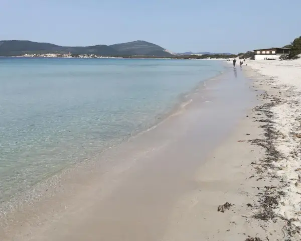 Spiaggia di Fertilia con acque cristalline, sabbia chiara e montagne sullo sfondo. Alcune persone passeggiano lungo la riva.