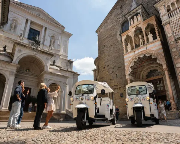 Tuk-tuk turistici pronti per esplorare cosa vedere a Bergamo in un giorno. Turisti scattano foto davanti a splendidi edifici storici.
