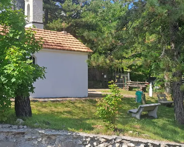 La piccola baita bianca, con tetto in tegole rosse, è circondata da alberi verdi e un campanile. Panchine di cemento invitano al riposo nel prato soleggiato.