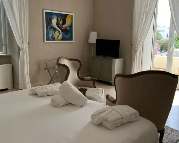 Camera accogliente al relais Corte degli Aragonesi, con letto, accappatoi, poltrone e vista su un balcone fiorito.