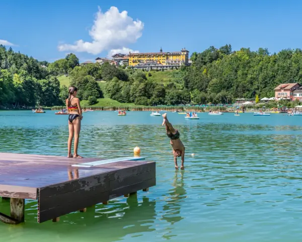 Ragazzo si tuffa in un lago balneabile, mentre una ragazza osserva dal pontile. Altri bagnanti e pedalò popolano le acque.