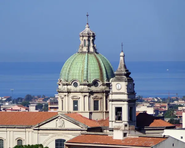 Cupola verde e campanile con orologio a Barcellona Pozzo di Gotto, con il mare azzurro sullo sfondo.