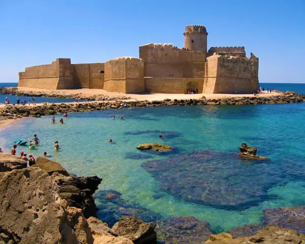 Castello Aragonese sull'acqua cristallina della costa ionica Calabria, con bagnanti che si godono il sole e il mare.