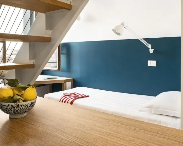 Un letto singolo con lenzuola a righe e un cuscino bianco, sotto una scala in legno e metallo. Un ciotola di limoni sul tavolo crea un'atmosfera da combo Venezia.