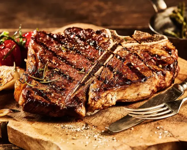 Una succulenta fiorentina, cotta alla perfezione, riposa su un tagliere di legno, pronta per essere gustata. Accanto, pomodorini e aglio arrostiti.
