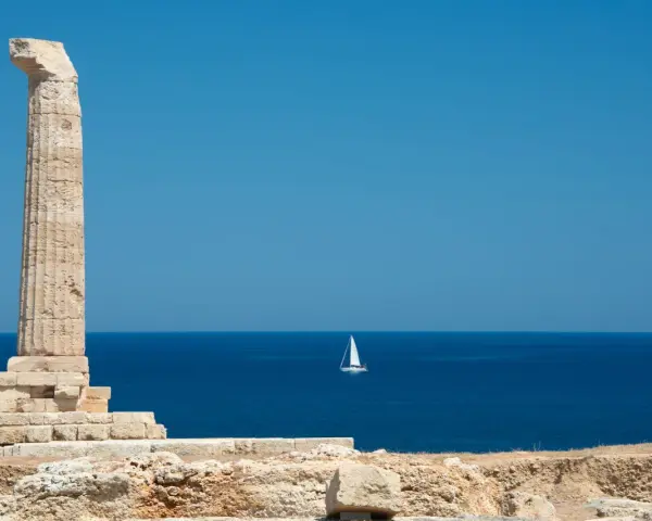 Antica colonna sulla costa degli Achei, con un veliero solitario all'orizzonte sotto un cielo azzurro.