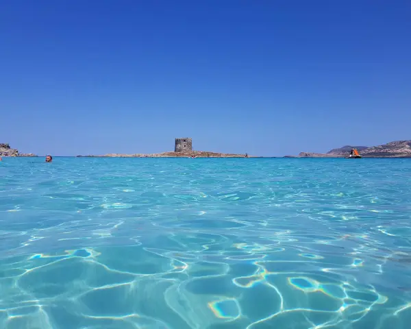 Acque cristalline e un antico torrione su un'isola: un mare bello in Italia, con gente che nuota e una barca a vela all'orizzonte.