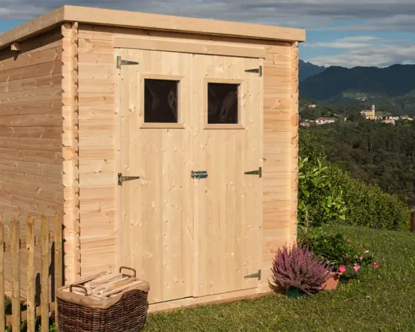 La casetta in campagna, in legno chiaro, con due finestrelle e un cesto di legna, si affaccia su un panorama collinare con un borgo in lontananza.