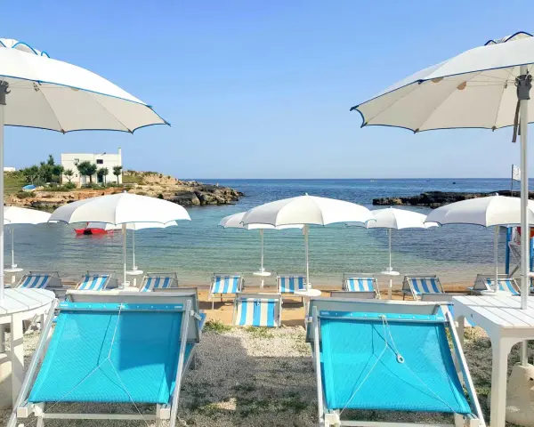 Lido Cala Pescatore: lettini azzurri e ombrelloni bianchi su una spiaggia di ciottoli, con il mare cristallino e rocce sullo sfondo.