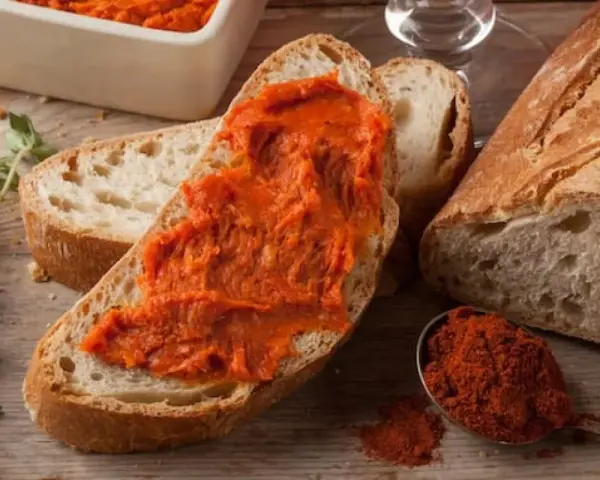 Fetta di pane con 'nduja, uno dei piatti tipici della Calabria, accanto a una ciotola con la crema e un cucchiaino di peperoncino in polvere.