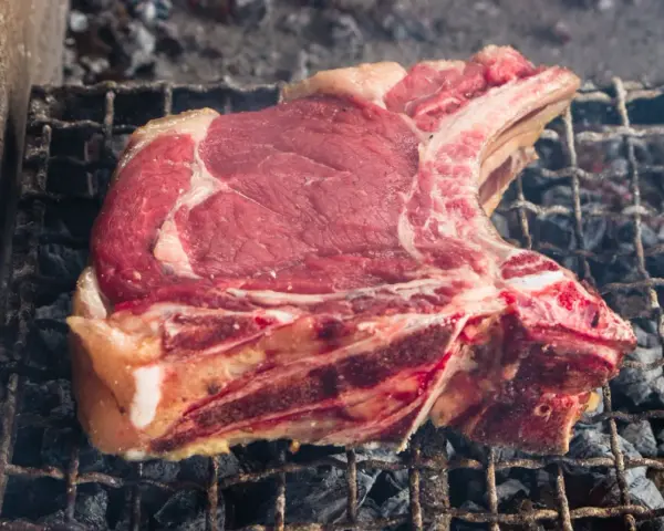 Una succulenta fiorentina di vitello, con la sua succosa carne rossa e il grasso invitante, cuoce lentamente sulla griglia ardente, sprigionando un profumo delizioso.