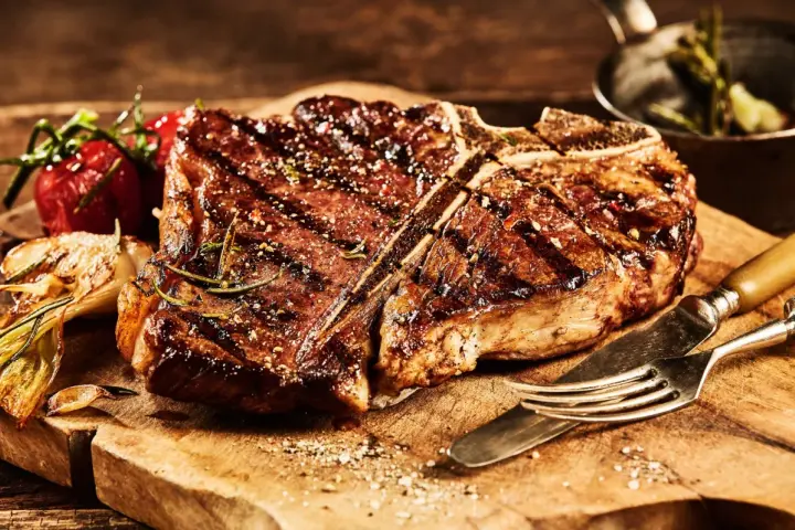 Una succulenta fiorentina, cotta alla perfezione, riposa su un tagliere di legno, pronta per essere gustata. Accanto, pomodorini e aglio arrostiti.
