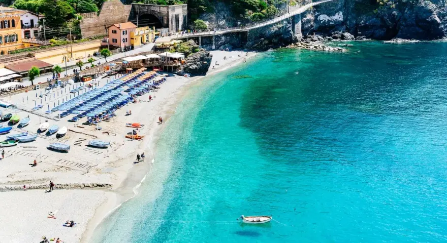 Spiaggia affollata con ombrelloni e lettini, barche sulla sabbia e mare cristallino. Un sentiero panoramico corre lungo le scogliere delle coste liguri.