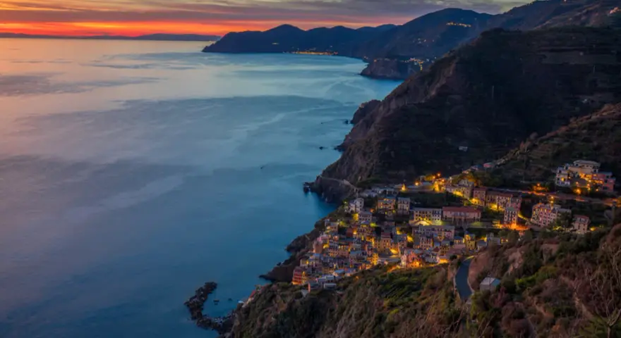 Riomaggiore cosa vedere: un villaggio colorato aggrappato alla scogliera, illuminato al tramonto, con il mare blu e le montagne sullo sfondo.