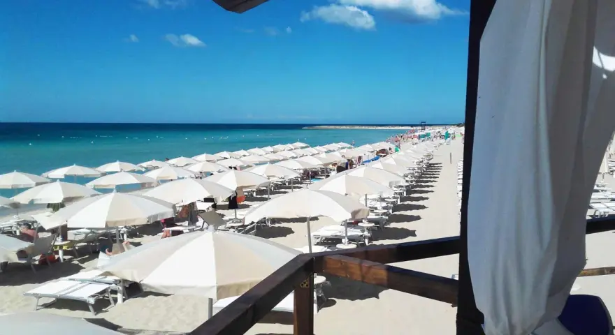 Ombrelloni bianchi e lettini ordinati sulla sabbia dorata, con il mare azzurro e il cielo limpido a fare da sfondo. Un'immagine perfetta di mamire lido Marini.
