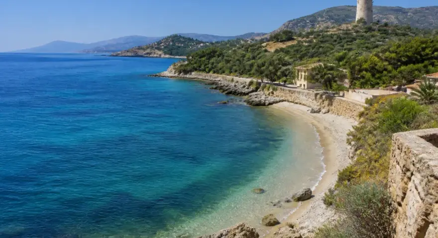 Spiaggia di Guidaloca con acque cristalline, vegetazione mediterranea e una torre antica in collina.