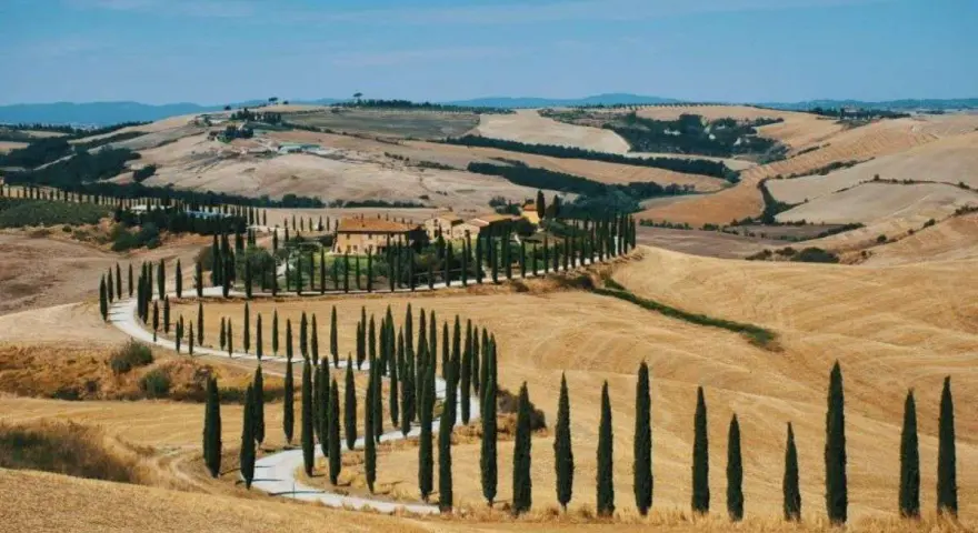 Un casale toscano circondato da cipressi lungo le strade del vino toscana, con dolci colline dorate sotto un cielo azzurro.