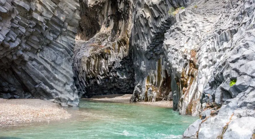 Acque cristalline scorrono tra le imponenti pareti basaltiche delle gole dell'Alcantara, creando una spiaggia naturale suggestiva.