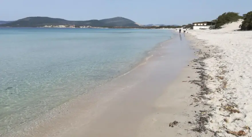 Spiaggia di Fertilia con acque cristalline, sabbia chiara e montagne sullo sfondo. Alcune persone passeggiano lungo la riva.