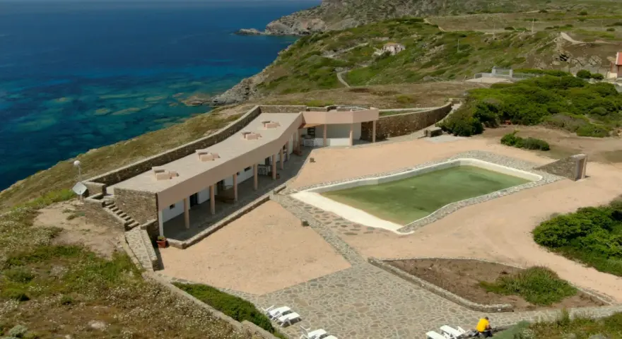 Un villaggio nurra affacciato sul mare turchese, con una piscina e lettini bianchi pronti per il relax.