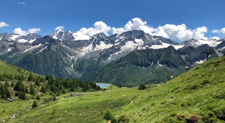 Un sentiero serpeggia tra prati verdi verso un lago alpino, con maestose montagne innevate sullo sfondo. Sembra il luogo ideale per una visita al villaggio delle marmotte.