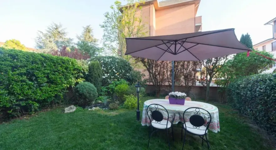 Un giardino curato con tavolo, sedie e ombrellone, circondato da siepi e alberi. Sembra l'albero dei gufi, un luogo magico per rilassarsi.