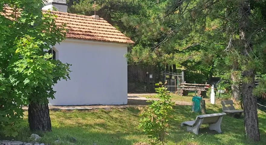 La piccola baita bianca, con tetto in tegole rosse, è circondata da alberi verdi e un campanile. Panchine di cemento invitano al riposo nel prato soleggiato.