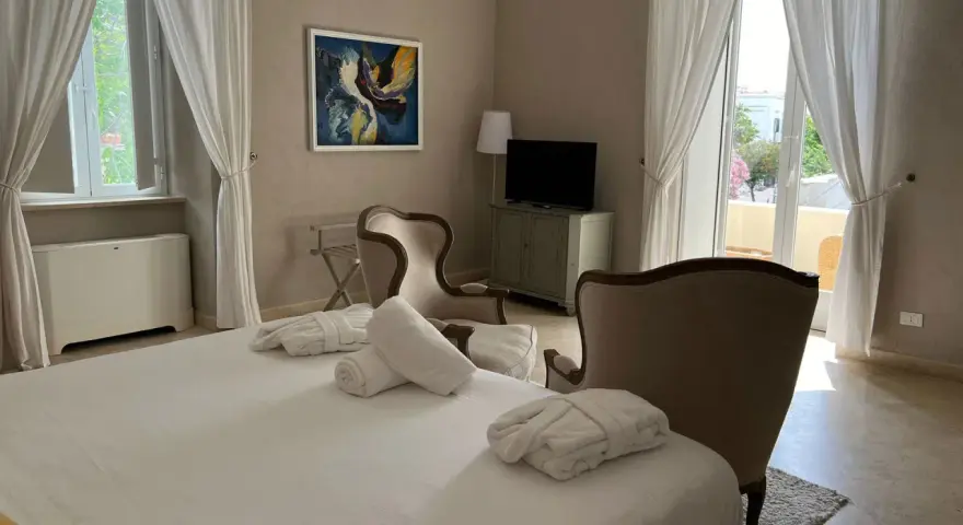 Camera accogliente al relais Corte degli Aragonesi, con letto, accappatoi, poltrone e vista su un balcone fiorito.
