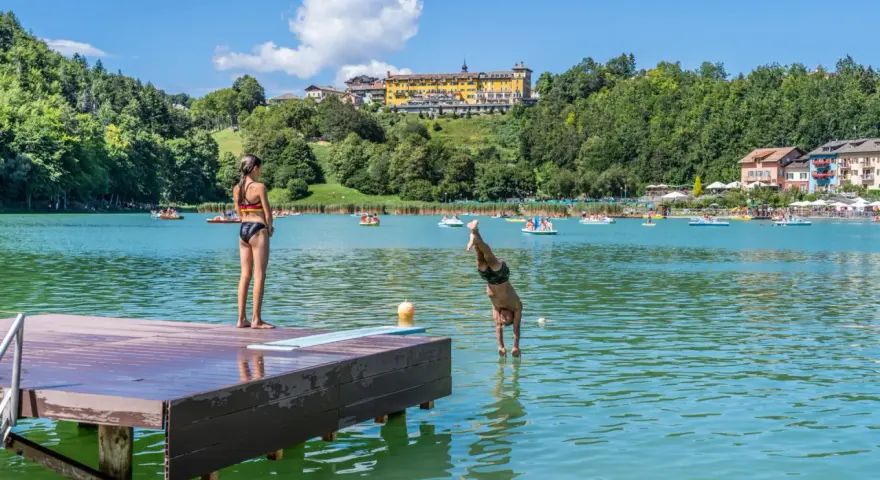 Ragazzo si tuffa in un lago balneabile, mentre una ragazza osserva dal pontile. Altri bagnanti e pedalò popolano le acque.