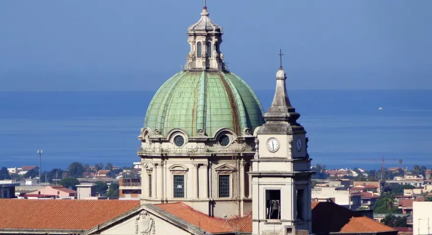 Cupola verde e campanile con orologio a Barcellona Pozzo di Gotto, con il mare azzurro sullo sfondo.