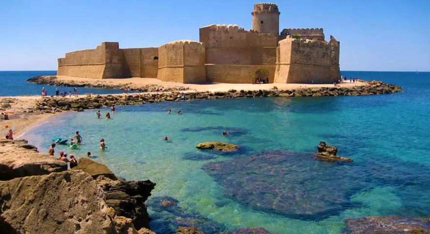 Castello Aragonese sull'acqua cristallina della costa ionica Calabria, con bagnanti che si godono il sole e il mare.