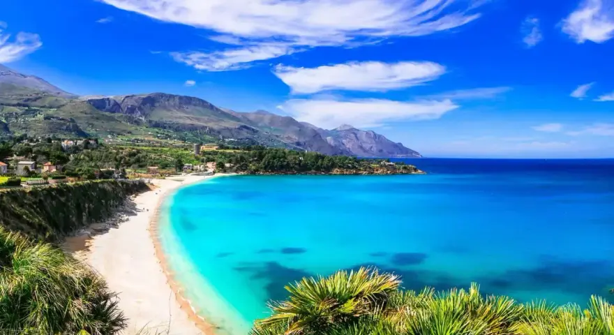 Una baia incantevole con acque cristalline e sabbia dorata, tra le spiagge più belle in Sicilia. Montagne verdi sullo sfondo.