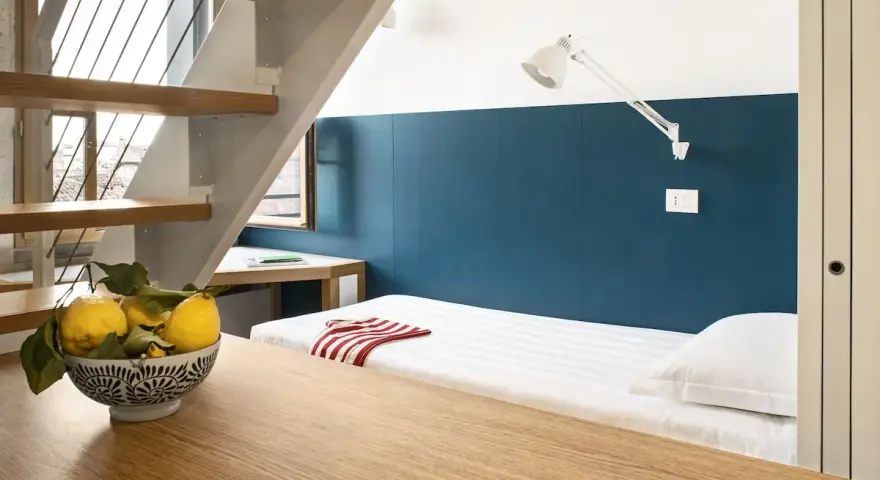 Un letto singolo con lenzuola a righe e un cuscino bianco, sotto una scala in legno e metallo. Un ciotola di limoni sul tavolo crea un'atmosfera da combo Venezia.