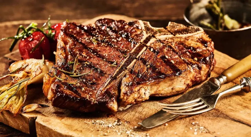 Una succulenta fiorentina, cotta alla perfezione, riposa su un tagliere di legno, pronta per essere gustata. Accanto, pomodorini e aglio arrostiti.